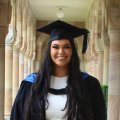 A woman with long black hair is smiling at the camera, she is wearing a graduation cap and gown over a white dress and is standing in the sandstone cloisters of UQ's Great Court.